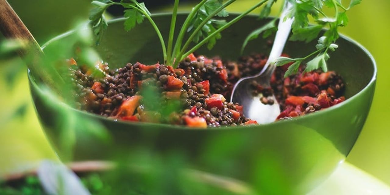 A green bowl filled with cooked brown lentils and chunks of carrot. The bowl is garnished with a small bunch of parsley and a teaspoon is resting inside.